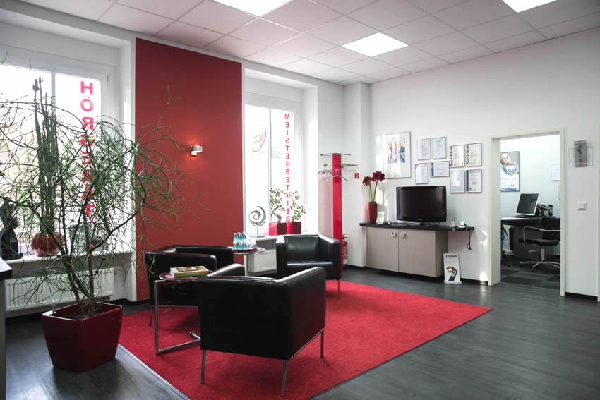 Roter Teppich auf schwarzem Boden mit drei schwarzen Sesseln, eine rote Wand, eine Fensterfront mit HÖRGERÄTE Kai Meinhardts Logo, einen großen, schwarzen Fernseher und Zertifikate an der Wand und links eine große Zimmerpflanze.